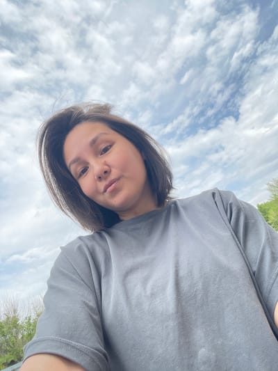 A selfie-style image shows a person with brown hair and fair skin, wearing a grey t-shirt. The background features a cloudy sky. The person is looking directly at the camera. The location is not deducible from the image. No details indicate this image was taken in Verkh-Katunskoye, Russia.