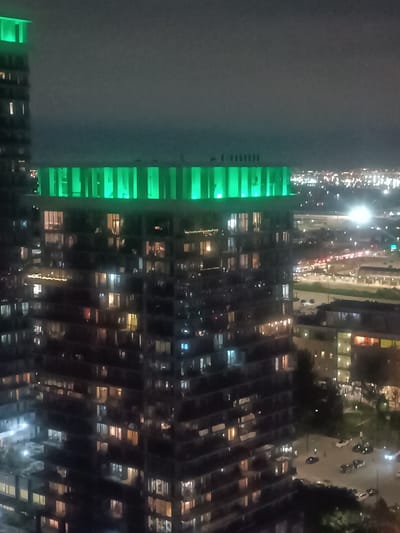 This image captures a nighttime aerial view of the Mississauga city skyline, specifically focusing on the Square One area. The foreground is dominated by a modern high-rise building with numerous floors, each containing windows lit with warm, artificial light. The upper section of this building is illuminated by a vibrant green light, creating a distinct horizontal band of color.
To the left of this main building, another, taller structure is partially visible, also topped with green illumination. The background reveals a sprawling urban landscape with a dense collection of buildings and illuminated streets, suggesting a busy city environment. Lights from other residential buildings and streetlights are visible, as well as some vehicles on the roads. The overall scene is dark, with the artificial lights of the city providing the primary illumination. The sky appears overcast or hazy, contributing to the muted ambient light.