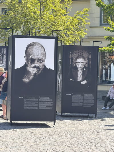 The image depicts an outdoor exhibition on a paved surface, likely a public square in the city of Lund, Sweden, given the presence of the ARKEN sign in the background. Two large black display panels are prominently featured. The left panel showcases a black and white portrait of Quincy Jones, a renowned musician and producer, with text below. The right panel displays a portrait of Ruth Bader Ginsburg, an Associate Justice of the Supreme Court of the United States, also with accompanying text. In the foreground, people are partially visible, with a woman wearing a straw hat and a patterned scarf on the left, and a man seated on a bench on the right. Trees with vibrant green foliage are present in the background, casting shadows on the ground. The overall scene suggests a cultural event or exhibition taking place on a sunny day.