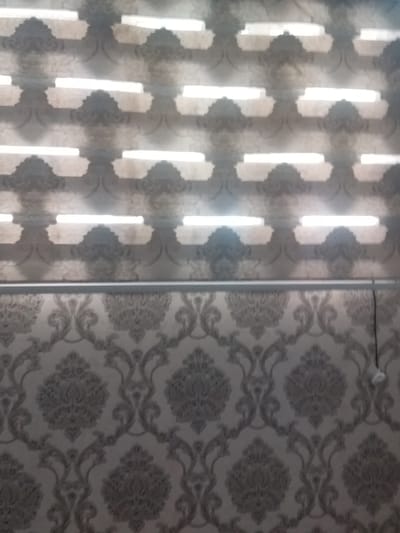 The image shows a patterned wallpaper. The upper portion of the wallpaper has horizontal bands of light and a repeating pattern of dark grey crests against a lighter background. The lower portion of the wallpaper features a more intricate damask pattern in dark grey against a lighter beige background. A thin, horizontal metal rod or bar runs across the image, separating the two distinct wallpaper patterns. A small white electrical outlet or light fixture is visible on the right side of the lower wallpaper section. The lighting in the upper portion suggests the presence of illuminated blinds or a similar window covering. The scene is indoors, likely a room with wallpapered walls. There are no people or explicit actions depicted.