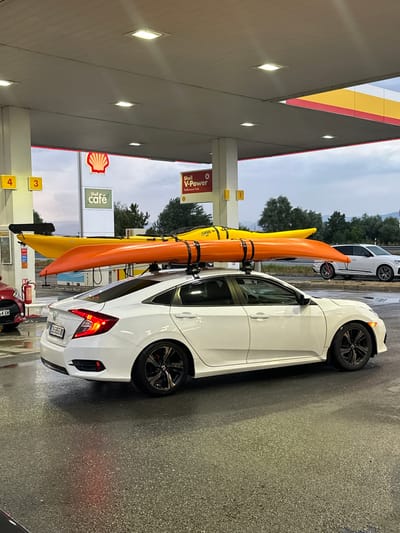 In Drazhevo, Bulgaria, at a Shell gas station, a white sedan is parked, its roof equipped with a rack carrying two kayaks, one orange and one yellow. The car's hazard lights are illuminated. Nearby, a red car is also parked. The gas station features Shell branding, including a large Shell logo, and signage for "Shell V-Power" and "Shell café". The ground is wet, indicating recent rain. In the background, trees and a cloudy sky are visible.