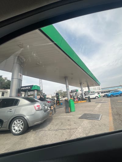 At a PEMEX gas station, a silver car is parked at a pump. Other vehicles wait in line for service. The gas station has a green roof and multiple pumps. A gray, metal plate is on the concrete pavement. The image is taken from inside another vehicle. Overcast skies are visible.