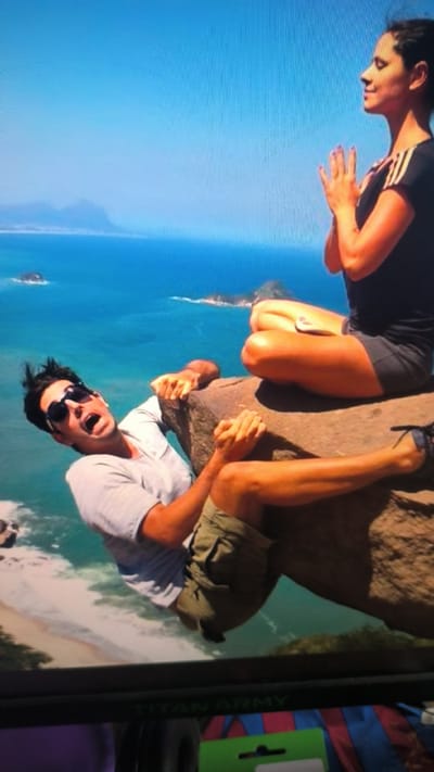 The image shows two people on a rocky cliff overlooking the ocean. On the right, a woman in a black t-shirt and shorts is meditating with her hands clasped in front of her chest. She is sitting cross-legged on the rock, facing away from the camera. On the left, a man in a light-colored t-shirt and khaki shorts is hanging precariously from the edge of the rock, with his mouth open in surprise or fear. He is looking towards the camera, and his sunglasses are perched on his nose. His hands are gripping the rock tightly. The ocean below is a vibrant blue, and there are islands or landmasses visible in the distance. The sky is a clear, bright blue, suggesting a sunny day. The overall scene depicts a daring and possibly humorous situation, with the contrast between the woman's calm meditation and the man's apparent distress. There is no visible text within the image itself.