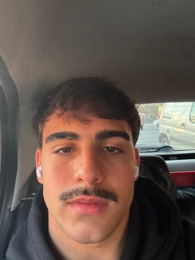 A medium shot of a young man's face in the interior of a vehicle. He has dark hair, thick eyebrows, and a trimmed mustache. He is wearing a black hoodie and has a white wireless earbud in each ear. Light is coming from the outside, indicating it is daytime. Reflections of other vehicles can be seen through the window. The presence of vehicles and surrounding structures suggests an urban setting. Without specific landmarks, determining the exact location within Quilmes Oeste, Argentina is not possible from this image alone.
