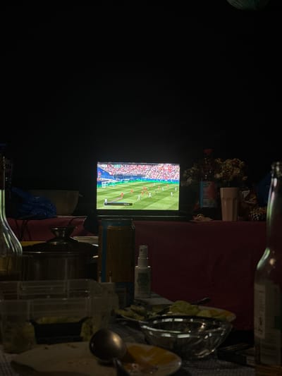 The image shows a television screen displaying a soccer match. The screen is positioned on a table, which is covered with a reddish tablecloth. Various objects are scattered on the table, including a glass container, a bottle, and some plates with food. The scene appears to be indoors, with the room dimly lit, suggesting it is nighttime. The background is dark, with some blurry elements that are not clearly identifiable. The focus is on the television screen, which shows players in red and white uniforms on a green field, with a large crowd in the stands. There is text on the screen, indicating "10.5 m". The overall impression is of a casual setting, possibly a living room or a communal area, where people are watching a sporting event. The lack of clear facial expressions or interactions makes it difficult to determine specific emotions, but the act of watching a game suggests engagement and perhaps excitement. There are also some dried flowers and a plastic bag visible on the table.