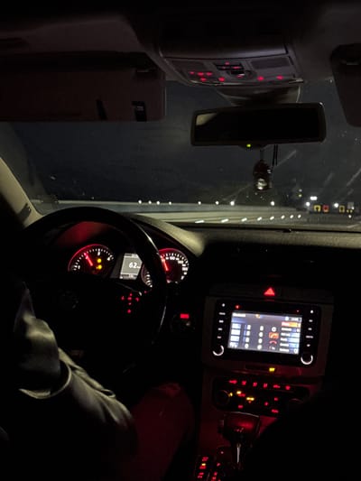 An interior shot of a car at night, taken from the passenger side looking towards the driver's seat and the road ahead. The overall scene is dark, illuminated primarily by the red and white lights of the car's dashboard and infotainment system.
In the foreground, part of a person's leg or arm, clad in dark clothing, is visible to the right. The steering wheel occupies the center-left, with the instrument cluster visible through it. The speedometer shows "62", likely indicating the vehicle's speed in kilometers per hour, alongside other markings like "140" and "180". A digital display within the cluster shows "21:19" (time) and "7.0". Red warning lights and indicators are active on the dashboard.
To the right of the steering wheel, the central console features an infotainment screen displaying a phone interface with options like "Phone", "Contacts", "Call Log", "Bluetooth", and "Settings". A time of "14:08" is shown on this screen, and icons for signal strength and battery are visible. Below the screen, the climate control panel shows illuminated buttons with text such as "AUTO", "AC", "OFF", and "DUAL". A hazard light button (red triangle) is also illuminated. Further right, a key can be seen in the ignition.
An overhead console with red illuminated buttons is visible in the upper left, next to the rearview mirror. Outside the front windshield, a dark environment with distant, scattered white lights suggests the car is either on a road at night or parked in a dimly lit area, possibly in Barakovo, Bulgaria. The rearview mirror shows a faint green light.