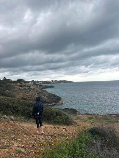A vertical photograph depicts a person standing on a rocky, dirt path beside a cliff overlooking the sea. The person is wearing dark clothing and a backpack, with pink shoes visible. The path is sparsely vegetated with low-lying plants and scattered yellow flowers.

The sea extends to the horizon with a slightly turquoise hue. In the distance along the shoreline, there are buildings and rocky outcroppings. The sky is overcast with long, striated clouds.

This image appears to be taken in Llucmajor, Spain.