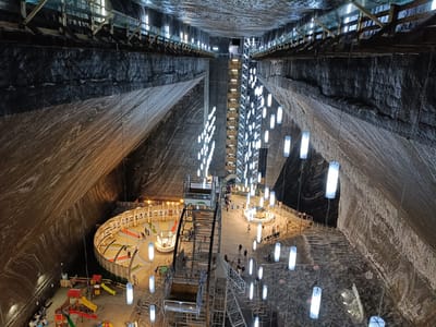 The image depicts the interior of the Turda Salt Mine in Turda, Romania, a former salt mine transformed into a tourist attraction. The vast cavernous space is illuminated by numerous hanging white cylindrical lights, creating a surreal ambiance. The walls of the mine are textured with layers of salt deposits, displaying a range of earthy colors from deep brown to off-white, with distinct striations.
In the foreground and mid-ground, various recreational activities are visible. A children's playground area with colorful slides, swings, and climbing structures is set up on a sandy floor. Adjacent to it, a miniature golf course with red and green turf winding through a circular area, featuring small decorative elements, is in operation with several people playing.
Dominating the central part of the mine is a large, industrial-looking elevator or vertical transport system, comprised of metal scaffolding and a cage-like structure. This system appears to connect different levels within the mine. Wooden walkways and railings are installed along the sides of the cavern, providing access to various vantage points and attractions.
People are scattered throughout the scene, engaged in activities like playing mini-golf, supervising children, walking on the pathways, and queuing for the elevator. The overall impression is a harmonious blend of natural geological formations and human-made infrastructure for entertainment. The lighting from the lamps casts a soft glow on the salt walls, highlighting their unique texture and vastness. The roof of the mine is visible at the top, with supporting wooden structures and more lights, suggesting it has been reinforced and adapted for public use.