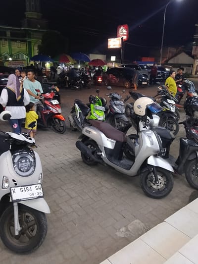The image captures a nocturnal scene in Sumberejo, Indonesia, depicting a bustling outdoor paved area, likely a parking lot or plaza, bordered by commercial establishments and a street.

In the immediate foreground, a light beige Honda Scoopy scooter (license plate K 6494 SW, valid until 06-30) is parked, facing forward. To its right, a silver Yamaha Fazzio scooter with a brown seat (partially visible license plate ending 5124 WR) is parked, displaying a beige helmet on its handlebars. The ground consists of grey interlocking concrete pavers.

In the mid-ground, a group of individuals and additional motorbikes are present. A man in a light blue t-shirt sits astride a red Honda Vario scooter (partially visible license plate beginning B #### U). To his left stands a woman wearing a white top, dark vest, and purple hijab, holding a white helmet. A young child in a yellow t-shirt stands next to the woman, facing the man on the scooter. Various other scooters, including green, black, and grey models, are parked throughout this section. To the far right, a woman in a yellow shirt and dark pants stands near a cluster of parked scooters, facing right.

The background reveals a street or open thoroughfare with several parked automobiles, including a dark hatchback and a white SUV. Multiple street vendor stalls operate under colorful umbrellas (blue, red, multi-colored), suggesting commercial activity. To the far left background, a green building featuring a minaret-like structure and a dome, characteristic of a mosque, is visible. To the right, a prominent, illuminated red sign for "Hits Chicken" is mounted on a commercial building. The entire scene is artificially lit by streetlights, building lights, and vehicle headlamps, indicating nighttime conditions.