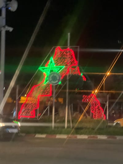 The image depicts an illuminated display of the map of Morocco at night, located in Beni Mellal, Morocco. The map outline is rendered in red LED lights, encompassing the full territorial claim of Morocco, including the Western Sahara. A large, five-pointed green star, also made of LED lights, is centrally positioned within the northern part of the map. Partially obscured behind the green star is a circular blue and white road sign, characteristic of a roundabout sign.
The display is supported by a metal framework, with several vertical posts visible behind it. In the foreground, a road is visible with a distinct red and white painted curb line running horizontally. A strip of green vegetation or grass is present between the curb and the light display. On the far left of the foreground, the front portion of a dark-colored vehicle with illuminated headlights is visible. On the far right, part of a yellow vehicle is discernible.
To the left of the main display, a tall utility pole is visible, equipped with multiple attachments that appear to be surveillance cameras or communication antennas. Diagonal light streaks and faint lines, possibly wires or reflections, traverse the upper portions of the image. The background is dark, indicating nighttime conditions, with distant, blurred lights suggesting other urban illumination. No individuals are visible in the scene. The overall image exhibits some blurring, particularly in the foreground and background elements, consistent with a low-light photograph.