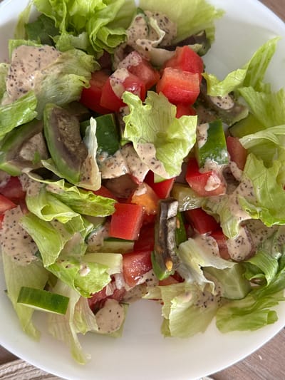 This is a close-up, top-down shot of a salad in a white bowl. The salad consists of chopped lettuce, tomatoes, cucumbers, and what appears to be avocado. The ingredients are coated in a creamy, speckled dressing. The background is a neutral, light-colored surface, possibly a table. There is no indication of the city of Sakmary, Russia, or any people present in the image.