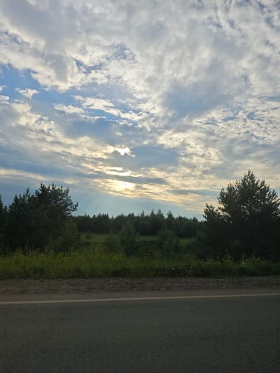 The image depicts a rural landscape with a road in the foreground. A dense line of trees, primarily pines and other coniferous varieties, forms a treeline across the middle ground. Beyond the treeline, a wide expanse of sky is visible, characterized by dramatic cloud formations. Rays of sunlight pierce through gaps in the clouds, creating a visually striking effect. The road is asphalt and appears to be in good condition, with a white line marking its edge. The presence of wild vegetation along the roadside suggests a natural, undeveloped environment. The scene is likely located in Povarenki, Russia, given the context provided. There are no people or animals visible in the image.