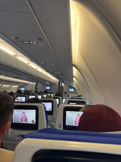 The image shows the interior of an airplane cabin, likely during flight. The cabin is filled with passengers seated in rows. Each seat has a personal entertainment screen displaying a video of a person in a pink shirt. Overhead, lighting illuminates the cabin. The scene appears to be in an airplane and no features indicate that the location is specifically Bol'shie Kabany, Russia.