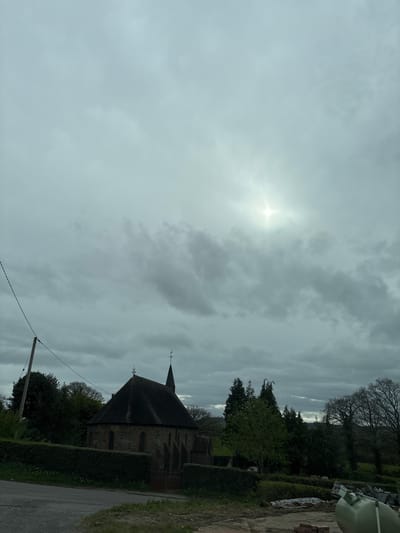This photograph, taken in Minsterley, United Kingdom, depicts a serene, overcast daytime landscape. The dominant feature is the cloudy sky, filled with thick, gray clouds through which the sun pierces, creating a bright, star-like flare in the upper-left portion of the frame.
In the midground, a traditional stone church with a dark, pointed roof and a spire or weather vane stands prominently on the right side. The church is bordered by dense green hedges and surrounded by a mix of trees; some exhibit bare branches, while others are in full leaf. A wooden utility pole with wires stretches across the lower part of the image.
A dirt or gravel road runs along the bottom-right edge of the frame. In the far upper-right corner, beyond the trees, hints of construction materials or debris are visible, including a large, light-colored object. No specific activity or people are present in the image. The weather is overcast but with moments of sun, suggesting a calm, quiet atmosphere. There is no visible text in the image.