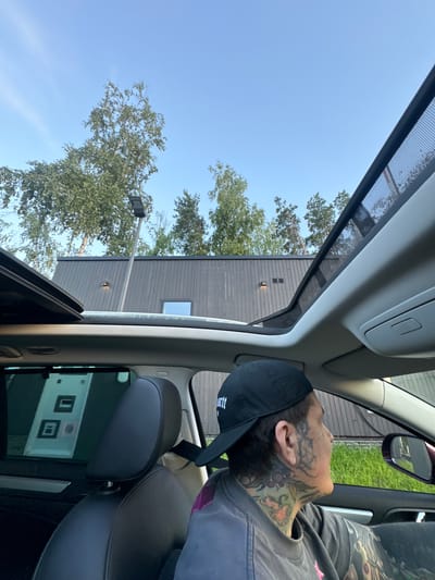 In the city of Barvikha, Russia, a person wearing a black baseball cap and a grey t-shirt is seen from the chest up, looking out the passenger side window of a car. The car's sunroof is open, revealing a clear blue sky with trees and a modern building in the background. The individual has extensive tattoos on their face, neck, and arm. The scene appears to be captured during daylight.
