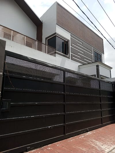 An exterior forensic image captures a modern multi-story residence in Viengthong, Laos, viewed from a street-level perspective on a partially overcast day. The property is secured by a large, dark-colored metal gate spanning the foreground, composed of solid horizontal panels on its lower two-thirds and a perforated metal section with small circular holes on its upper third. A smaller access panel is integrated into the left side of the main gate. The ground in front of the gate is paved with reddish-brown bricks.

The residence features white exterior walls accented with dark trim and decorative elements. On the left side of the upper floor, a balcony with silver-colored horizontal railings extends, adjacent to a section with a dark wood-like vertical panel under a pitched roofline. On the right, the building exhibits a flat roofline with a large, textured brown rectangular panel below it. Dark-tinted windows are present, with one window on the upper left appearing partially open. Below the right upper window, a decorative pattern of irregular dark grey rectangular panels is affixed to the white wall. A smaller, white, flat-roofed structure with dark trim is visible above the gate, suggesting an entrance or architectural feature. Multiple black utility lines are visible against the cloudy sky in the upper portion of the frame. No individuals or specific actions are depicted within the scene.