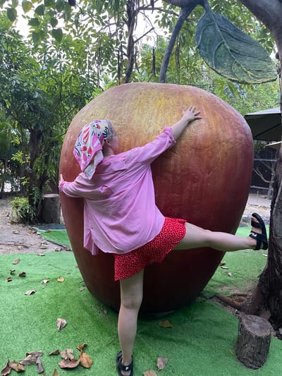 A rotated outdoor shot shows a woman wearing a pink shirt, red polka dot dress, and a patterned headscarf, posed against a giant artificial apple. She is leaning against the apple with one hand and leg extended in a partial split. Her feet are clad in black sandals. The apple is a mix of red and yellow hues, and its stem and leaves are visible. Artificial green turf covers the ground where the woman's feet rest, and some fallen leaves are scattered about. Trees and foliage frame the scene. The shot was taken at Nha Trang, Vietnam.