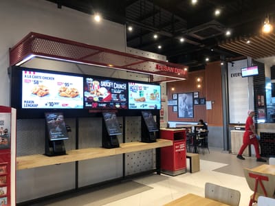 The image captures the interior of a brightly lit KFC fast-food restaurant in Triengmatangubi, Indonesia. The central focus is a long counter with multiple large digital menu boards displaying various fried chicken meals, drinks, and prices in Indonesian Rupiah. Text like "A LA CARTE CHICKEN," "SAUCIFY CRUNCH," and "CHAKIT MEAL" are visible on the screens, along with promotions such as "MAKIN LENGKAP" (more complete) and "JAGONYA RASA ORIGINAL" (expert of original taste).

Below the digital menus are several self-order kiosks, each with the text "KETUK UNTUK MULAI" (tap to start). The kiosks rest on a light wooden countertop, supported by a metal frame with a mesh panel. To the right of the kiosks is a red trash bin, featuring an illustration of a person disposing of waste and the text "Mulai #BUDAYA BEBERES DARI DIRI SENDIRI" (Start #CULTURE OF CLEANING UP AFTER YOURSELF).

In the background, a KFC employee in a red uniform, including an apron and head covering, walks towards the right side of the frame. Further back, a customer is seated at a table, engrossed in their phone. The restaurant features a mix of modern and rustic design elements, including wooden slat walls, white walls, and a black ceiling with exposed ventilation and recessed lighting. A pillar on the right lists the days of the week from "SUNDAY EVERY" to "SATURDAY." Other visible texts include "PESAN DISINI" (order here), "HERBS AND SPICES," "FINGER LICKIN GOOD," and "ESTD 1940." A disclaimer at the bottom of one screen indicates that listed prices do not include a 10% tax. The overall atmosphere is clean and functional, indicative of a typical fast-food dining establishment.