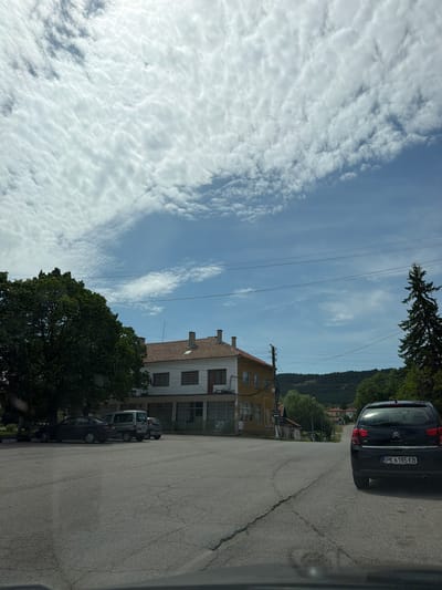The image shows a street scene in Stefanovo, Bulgaria. A gray Citroen car, with license plate PK 6185 KB, is parked on the right side of the road. A light yellow two-story building with a red tile roof and multiple windows is visible along the road. A large green tree is partially obscuring the building. Several other cars are parked further down the street, near the building. Power lines run across the sky. The sky is blue with scattered white clouds. The road is asphalt and shows signs of wear and cracking. A wooded hill rises in the background. The image appears to be taken from inside a vehicle.