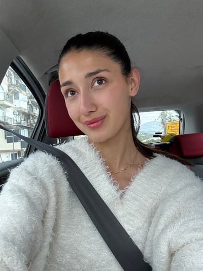 A woman with dark hair pulled back is in a car. She is wearing a white, fuzzy sweater and a gold chain necklace. A black seatbelt is across her body. The interior of the car includes a red headrest. Outside the car window, buildings are visible, with a yellow sign that shows directions to Ulcinj, Bar, and Sutomore. The location appears to be Zgrade, Montenegro.