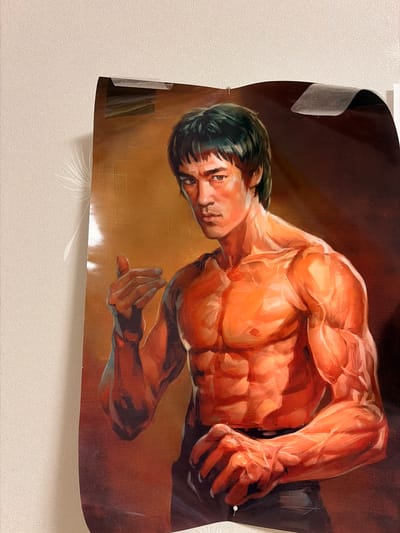 This is a medium shot, eye-level photograph of a poster of Bruce Lee, a martial artist and actor, hanging on a wall. The poster is a painting depicting Bruce Lee in a dynamic pose, showcasing his muscular physique. He has dark hair and a serious expression. The poster is attached to a textured, light-colored wall with clear tape at the top and a white thumbtack at the bottom. The lighting appears to be warm and directional, creating highlights and shadows on Bruce Lee's body and the poster's surface. The background of the poster is a gradient of warm tones, from a golden yellow to a dark brown. There is no visible indication of the city of Leninogorsk, Russia, in the image itself.