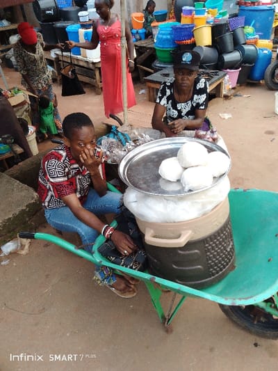 Two women sell food at an outdoor market. One woman in a red and white patterned shirt sits in a green wheelbarrow next to a cooler with a silver tray of white balls, her fingers near her mouth. Another woman wearing a black cap and patterned shirt sits behind the wheelbarrow with other food items. Various objects, including buckets, baskets, and people, fill the background, indicating a busy market setting.
