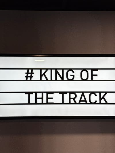 A close-up vertical shot depicts a backlit message board with black cutout letters set against a white illuminated background. The board displays the text "#KING OF THE TRACK" across two lines, with "#KING OF" on the upper line and "THE TRACK" on the lower line. A thin black frame encloses the illuminated white display area. Above the sign, a single, small, circular recessed light fixture is visible, casting illumination downwards onto the top edge of the board. The surrounding wall, both above and below the sign, appears dark brown or grey, with a subtle textured finish. No individuals are present. This scene is located within Eastleigh, United Kingdom.