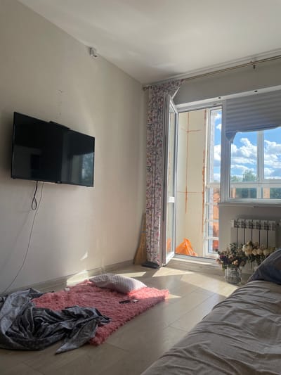 The image shows the interior of a room with a large window and a balcony door, likely located in Obninsk, Russia. Sunlight streams into the room, casting long shadows on the wooden floor. To the left, a black flat-screen television is mounted on a light-colored wall. Below the TV, a small pink rug is laid out, with a white pillow and a grey patterned blanket on top of it.
The window and balcony door are open, revealing a construction site on the balcony, indicated by orange insulation and an orange plastic bag. Outside the window, a clear blue sky with fluffy white clouds is visible, along with trees and buildings. A white radiator is positioned beneath the window, and a bouquet of dried flowers in a plastic bottle sits on the windowsill. In the foreground, the corner of a bed with grey and white patterned bedding is visible. The room appears to be a bedroom or living area.