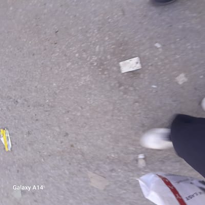 The image shows a section of pavement, likely in Al Mawasi, Unknown, given the metadata. A portion of a white shoe, black clothing, and a partially visible white bag with a red stripe and the text "EAST" are present in the lower right corner. There is some scattered debris on the pavement, including a small piece of yellow and white trash in the lower left, a small white rectangular object near the center, and a piece of cardboard near the bottom. The overall scene appears to be at ground level, with a focus on the surface and surrounding objects.