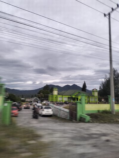 This media file captures a scene in Panteraya, Indonesia, likely during the day under overcast skies. The foreground is blurred due to motion, suggesting the image was taken from a moving vehicle. Several vehicles, including cars and a motorcycle, are visible in what appears to be a parking area or road. 

In the middle ground, a distinctive green building with yellow accents stands prominently. It has a symmetrical design with multiple windows and a tower-like structure on the right, topped with a red and white flag. The building's architecture suggests it could be a public institution, a school, or perhaps a place of worship. 

In the background, rolling hills and mountains are silhouetted against a cloudy sky. The overall atmosphere is somewhat muted due to the cloudy weather, but the bright green of the building provides a visual anchor. The presence of power lines overhead is a common sight in many populated areas and adds to the everyday urban or suburban setting. The motion blur in the foreground and the general lack of sharp detail prevent the identification of specific individuals or detailed emotional expressions.