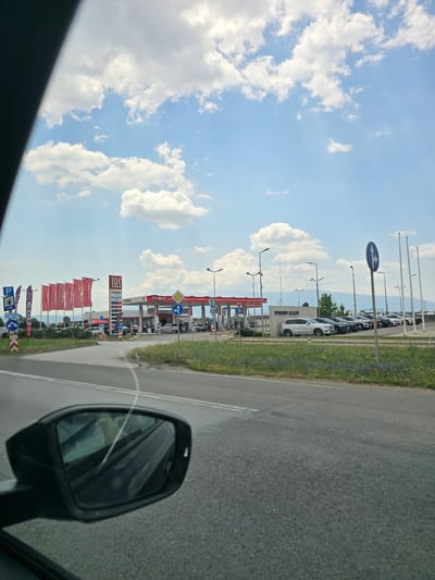 The image depicts a roadside scene in Krivina, Bulgaria, on a sunny day with scattered clouds. The perspective is from inside a vehicle, looking out towards a gas station and a row of parked cars. The gas station, identified by the "Lukoil" and "OES" logos, features red and white branding and several gas pumps. Red flags with white text are displayed along the roadside in front of the gas station.

Parked on the other side of the gas station are several cars, suggesting a car dealership or a display area. Road signs are visible, including directional arrows and a parking symbol. The road itself is paved with asphalt and has white lane markings. A grassy verge with wildflowers lines the side of the road. In the foreground, a car's side mirror is visible, reflecting the sky and some of the surrounding scene. The overall atmosphere is that of a typical day at a service area or along a highway in a rural or semi-urban setting.