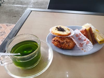 The image shows a close-up, overhead view of a table setting with food and drink. A clear glass cup filled with a vibrant green liquid, likely a beverage, sits on a matching saucer. A metal spoon rests inside the cup. To the right, on a light blue plate, are several small food items. These include what appears to be a small, round pastry topped with dark sprinkles, a golden-brown fried item, and two small sandwiches with pinkish filling, one of which is wrapped in white paper with a floral pattern. The table surface is a neutral beige color, and a dark-colored table frame is visible in the background, along with a portion of a tiled floor and what appears to be the edge of a chair. The scene suggests a casual meal or snack. No specific details about Palohlada, Indonesia are visible.