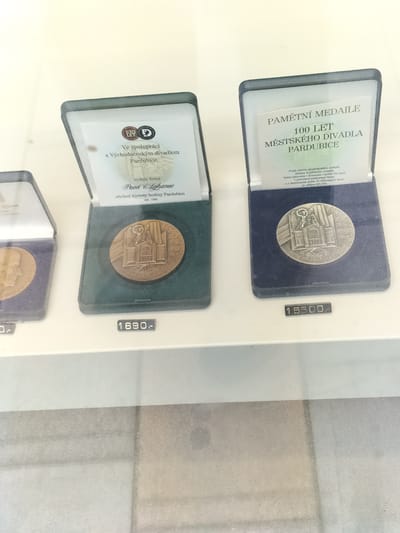 The image displays two display cases containing commemorative medals, likely in a museum or shop in Pardubice, Czechia. The left display case, partially visible, contains a medal in a dark green velvet-lined box. A label below it reads "0.-". The right display case, more prominently featured, contains a bronze medal within a dark blue velvet-lined box. The medal depicts a building with an angelic figure above it and text around the rim. A label above the medal reads "PAMĚTNÍ MEDAILE 100 LET MĚSTSKÉHO DIVADLA PARDUBICE", indicating it commemorates 100 years of the Pardubice City Theatre. Below this medal, a label reads "15500.-". A second, partially visible display case to the left of the main one shows a label below it reading "1690.-". The overall environment appears to be an indoor exhibition or retail space with a light-colored background.