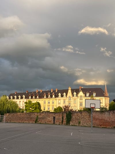 The image depicts an outdoor basketball court in front of a brick wall, with a building in the background. The court is asphalt with white markings. A basketball hoop stands on the right side of the image. The brick wall is weathered and covered in ivy in some areas. Behind the wall, a long, yellow building with multiple windows and a dark tiled roof is visible. The building has a spire on its right side and appears to be historical. The sky above is dramatic, with dark, heavy clouds mixed with patches of lighter, illuminated clouds, suggesting an impending storm or the aftermath of one. Lush green trees, including a weeping willow, frame the left side of the building and wall. The scene is set during what appears to be either early morning or late afternoon, given the golden light illuminating the building and clouds. The setting is likely Colmar, France, given the architectural style of the building. No people or actions are depicted.