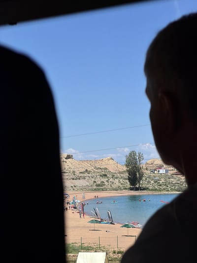 The image depicts a view of Lake Issyk-Kul, specifically a beach area near Tong, Kyrgyzstan, as seen from inside a vehicle. In the foreground, the silhouettes of two people's heads and shoulders obscure parts of the scene. The midground features a sandy beach populated with numerous people engaged in various activities. Several windsurfing boards and sails are propped up on the sand, and some individuals are in the water, some on paddleboards and others on what appear to be small boats. A green umbrella provides shade on the beach. The background shows a body of blue water, Lake Issyk-Kul, and beyond that, arid, hilly terrain characteristic of the region. A few trees and small buildings are visible on the distant hills. The sky is clear and blue.
