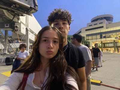 A group of people are gathered near a jet bridge at an airport in Ferno, Italy. In the foreground, a young woman is taking a selfie, with a young man partially visible behind her. The woman is wearing a light-colored, long-sleeved shirt and a necklace. The man is wearing a black t-shirt and a chain. In the middle ground, other individuals are standing, appearing to be waiting or boarding. To the left, there is a woman in a white t-shirt holding a dark bag. In the background, an airport terminal building with distinctive architecture and a control tower is visible. The sky is a dusky blue, suggesting it is evening or early morning. The ground is paved, and there are yellow safety markings and a jet bridge connecting to an unseen aircraft.