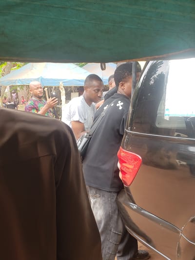 The image, taken in Umundugba, Nigeria, captures an outdoor gathering of several men during the daytime. The scene is framed by a green fabric awning on the left and the brown clothing of the person holding the camera in the right foreground.
In the mid-ground, a black car, likely a sedan or SUV, is parked, with its rear section, including a prominent red taillight and side windows, clearly visible. Two men are standing close to the car. One man, wearing a white short-sleeved t-shirt and a black shoulder bag, has short dark hair and faces towards the left of the frame. Behind him, another man in a black hoodie with a distinct white cross-like emblem on the chest and dark jeans is positioned near the car's rear passenger door. A person with a beard is partially visible near the front of the car, reflected in the window.
Further to the right, a bald man wearing a vibrant, multi-colored patterned shirt is actively gesturing with his right hand, suggesting he is speaking or explaining something to the group. Other individuals are faintly visible in the background, standing under what appears to be a light blue tarp or canopy, providing shade. The ground beneath them seems unpaved, consistent with an outdoor setting. The lighting is bright and natural, indicating it is daytime, possibly with an overcast sky given the lack of harsh shadows. The overall atmosphere suggests a casual community interaction or event. There is no discernible text visible within the frame.