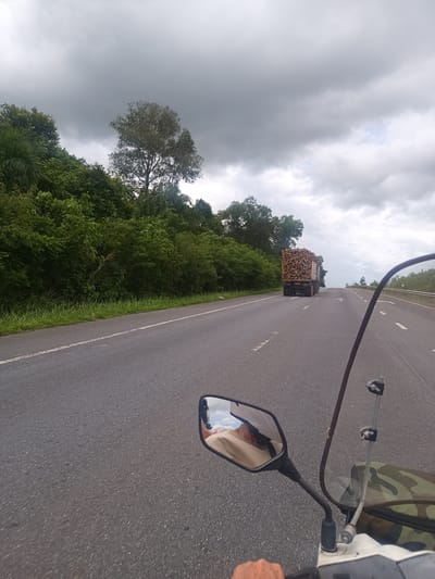 A view from a moving motorcycle on a multi-lane highway in Khian Sa, Thailand. The road, with white lane markers, stretches ahead with a gradual upward slope. A large flatbed truck filled with what appears to be wooden logs is in the distance, moving in the same direction. Lush green vegetation lines the left side of the road. Overcast skies create a muted, diffused light. In the foreground, the motorcycle's left side mirror reflects the driver and the sky. The motorcycle has a camouflage bag and a windscreen. The driver's hand is visible on the handlebars.