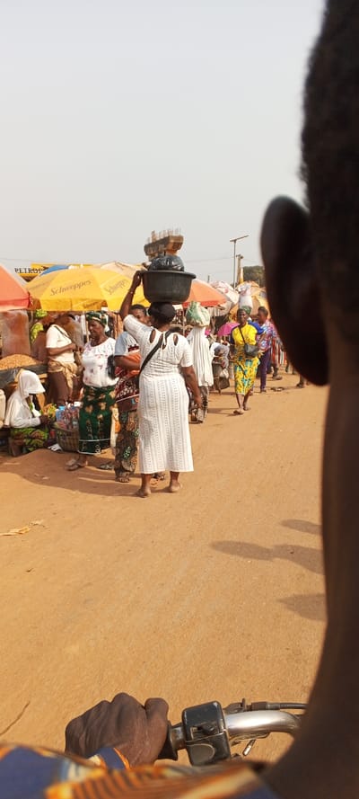 This image captures a bustling outdoor market scene in Ketu Adieowe, Nigeria, viewed from the first-person perspective of someone on a motorcycle, indicated by the visible hand on a handlebar and a shoulder in the foreground. The ground is a reddish-brown dirt road, characteristic of unpaved surfaces.

The scene is filled with numerous people, mostly women, engaged in daily market activities under bright daylight, suggested by the clear shadows on the ground. The sky appears bright and somewhat hazy. In the midground, a woman in a white dress stands prominently, expertly balancing a black basin filled with items, including a black plastic bag, on her head. Other women are seen walking, standing, and interacting within the market, many wearing colorful traditional attire and headwraps. Some vendors are seated beside their stalls.

The market features various stalls covered by colorful canopies. A yellow canopy prominently displays the brand name "Schweppes." Another canopy shows the text "PETROG," possibly part of a "PETROL" or "PETROGEN" sign. In the background, a large concrete structure with visible rebar indicates ongoing construction or infrastructure. The overall setting portrays a vibrant and active commercial hub, typical of a West African open-air market.