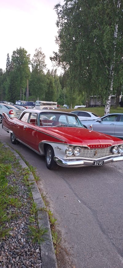 In Salpakangas, Finland, a vintage red and white Plymouth Fury sedan is parked on the right side of a paved road, adjacent to a gravel and grass verge. The car, which has chrome accents and a distinctive grill, faces towards the left side of the frame. In the background, a line of trees, including birches and other deciduous trees, creates a backdrop. Behind the trees, various vehicles are visible, including a white bus and a silver car, suggesting a parking lot or a road with other parked vehicles. The sky is a pale, overcast hue.