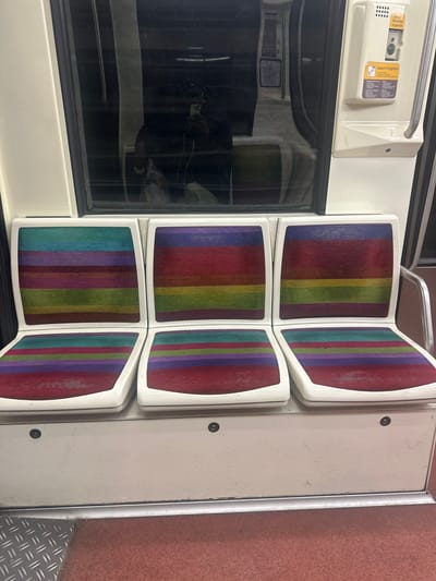 The media file captures the interior of what appears to be a public transport vehicle, likely a subway or train car, given the context of Neuilly-sur-Seine, France. The scene is dominated by a set of six fold-down seats, arranged in two vertical columns of three, against a white wall. Each seat cushion features a distinct vertically striped pattern in various colors including red, green, blue, purple, and yellow.

To the left of the seats, a dark window reflects a partial image of a person, presumably the photographer, holding a phone. Above the window, an emergency call panel is mounted on a light-colored wall. A silver metal handrail extends along the top of this panel.

The emergency panel features prominent text in French. The heading "Danger Sécurité Urgence" (Danger Safety Emergency) is visible at the top. Below, the phrase "Appel d'urgence" (Emergency call) is displayed, followed by instructions: "1. Tirer le capot" (1. Pull the cover), "2. Appuyer sur le bouton" (2. Press the button), "3. Attendre la réponse" (3. Wait for the answer), and "4. Décrivez la situation" (4. Describe the situation). A small icon of a phone handset is also visible.

The floor to the right of the seats is a reddish-brown, possibly carpeted or textured surface, transitioning to a metallic section with a diamond-plate pattern. The interior appears to be under artificial lighting, and while generally clean, some minor wear and scratches are visible on the white panels. The overall atmosphere is functional and indicative of public transportation.