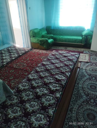 The image depicts the interior of a room, likely a living room or bedroom, with a focus on the floor and furniture. The room is decorated with several rugs, featuring intricate floral patterns in shades of red, maroon, and cream. A green sofa with a textured pattern is visible in the background, positioned in front of a bright window covered by turquoise curtains. The floor is covered with multiple rugs layered on top of each other, some appearing to be bedding or floor mats with a maroon base and a repeating cream and white floral design. There's a hint of a doorway on the left, leading to an outdoor area with light filtering through. A wooden-topped object, possibly a low table or bench, is present in the foreground, partially obscuring the rugs. The overall impression is of a well-used, comfortable living space. The date stamp on the image indicates "30.07.2026 12:51". The location is stated to be Quyosh, Uzbekistan.