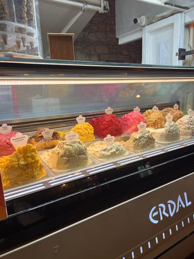 The image depicts an illuminated ice cream display case inside a commercial establishment in Çeşme, Turkey. The foreground features two rows of large, square metal tubs containing different flavors of dondurma or gelato. Each flavor is identified by a small, white, heart-shaped label, typically listing the name in both Turkish and English. Visible flavors include Visne Cherry, Mandarin, Kahve Coffee, Cici Bebe ingilüxe, Mango, Frambuaz (raspberry), Big Babol, Cevizli Ceviz (walnut), Kestane (chestnut), Keci Sutu Goat Milk, Sade (plain), Kurabiye (cookie), and Tutti Frutti. The ice cream displays a range of colors, including various shades of red, pink, yellow, brown, and white, with different textures. The lower panel of the display case is marked with the illuminated word "ERDAL" in a stylized font, along with a row of illuminated vertical dashes. Above the display case, to the upper left, a clear plastic cylindrical container holds several small, conical waffle cones. The background behind the display features a textured stone wall, a white wooden door or frame, and a white security camera mounted on a white beam above the frame, pointing downwards and left. No individuals or active interactions are present in the scene.