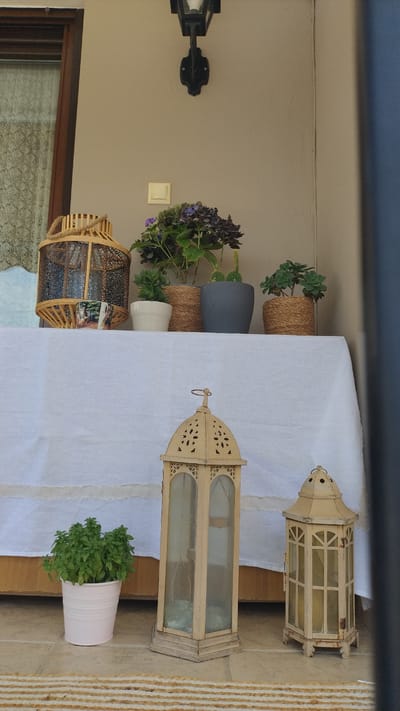 The image depicts an outdoor scene, possibly a porch or balcony, in Νέα Μηχανιώνα, Greece. A table draped with a white linen cloth is set up. On the table, there are several potted plants, including a hydrangea with purple blooms, succulents, and a cactus. Decorative elements include a rustic wooden lantern and a mug with a landscape scene. In front of the table, on the floor, stand two ornate, aged metal lanterns, one tall and rectangular, the other hexagonal and shorter. A small potted basil plant is placed to the left of the lanterns. The background shows a beige-colored wall with a black outdoor light fixture mounted above. The floor appears to be paved with light-colored tiles, and a striped rug or runner is visible in the foreground. The overall impression is one of a decorated outdoor living space.