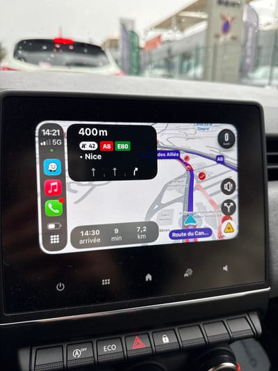 The image depicts the interior of a vehicle, primarily focusing on its infotainment screen, with a blurred view of the exterior through the windshield. The infotainment screen displays Apple CarPlay navigation, indicating the vehicle's location in Mougins, France, near the intersection of roads like "Route des Alliés" and "Route du Cannet," close to businesses such as "Renault Retail Group" and "Volkswagen Vehicules Utilitaires Riviera Technic SA."

The navigation shows a current instruction to proceed 400 meters towards "Nice" via routes N 42, A8, and E80, with an estimated arrival time of 14:30, 9 minutes, and 7.2 kilometers. The current time is 14:21, and the vehicle is stationary (0 km/h). Raindrops are visible on the windshield, suggesting overcast or rainy weather. Beyond the windshield, blurred commercial buildings and a portion of a white car with a red brake light are visible. Below the infotainment screen, physical vehicle control buttons are present, including "A OFF" (Start/Stop disable), "ECO," and a hazard light switch. No individuals are visible in the scene.