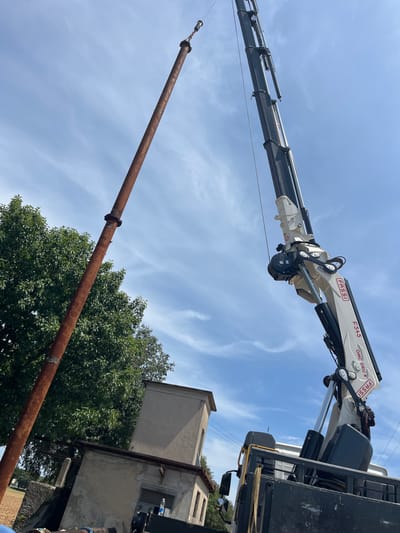 In the city of Castiglione delle Stiviere, Italy, under a partly cloudy blue sky, a long, rusty metal pipe is being lifted vertically by a crane. The crane, a Fassi F545 OMC GRU, is attached to the back of a truck. The pipe, approximately 10-15 meters long and 30 centimeters in diameter, is suspended from a hook at the end of the crane's boom. The pipe appears to be made of steel and shows signs of corrosion. In the background, there are some buildings and trees. The buildings are low-rise and appear to be industrial or utility structures. The trees are green and leafy. The overall scene suggests construction or demolition work is taking place.