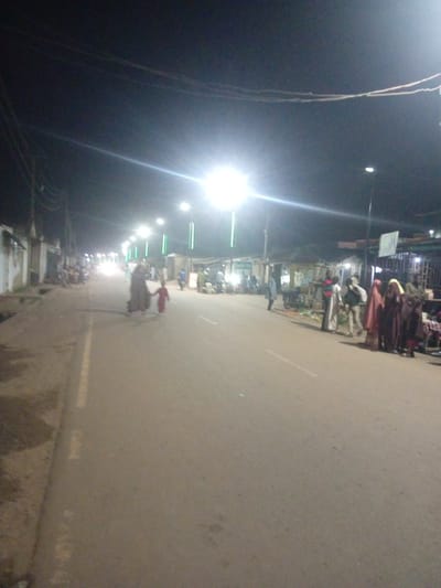 The image depicts a street scene at night in Tundun Wada West, Nigeria, illuminated by streetlights and possibly some ambient light from buildings. The road is paved and appears to be a two-lane street. On the left side of the road, there are buildings with what looks like a wall or fence. On the right side of the road, there are more buildings, some appearing to be shops or market stalls, and a variety of people are visible.

In the foreground, a woman in a dark outfit is walking with a young child dressed in red. They are walking on the road towards the viewer's left.

Further down the street, on the right side, a group of people are gathered, some standing and some possibly seated. They are near what appears to be stalls or displays, suggesting a market or roadside commerce is taking place. Several individuals are wearing traditional attire, including headscarves and long garments. Some people are interacting with each other, and others are looking towards the stalls. There are also what appear to be motorcycles parked in the area.

The streetlights are prominently featured, casting bright illumination and creating a somewhat hazy atmosphere due to the night and potential for dust or mist. Electrical wires are visible overhead, strung between poles. The overall impression is of a busy street at night, with people engaged in various activities, likely related to a market or evening social gathering.