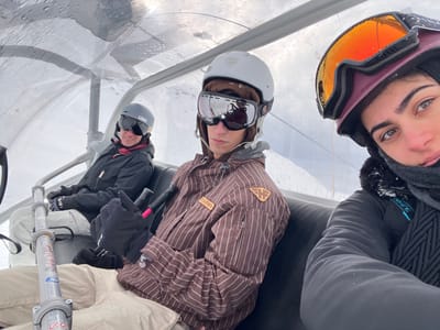 Three individuals are seated from right to left on a covered ski lift chair in Valtournenche, Italy. The chair features individual dark padded seats and is enclosed by a clear plastic bubble canopy, which has visible water droplets on its surface.
The person on the far right, partially visible in the foreground, is a female wearing a dark purple ski helmet with orange-tinted ski goggles pushed up, revealing her eyes. She wears a dark jacket with snow specks and a dark neck scarf. Her expression is neutral.
To her left, in the middle of the frame, is a male individual. He wears a light grey ski helmet with silver-mirrored ski goggles, a brown and white pinstriped ski jacket with a light brown "FEATHER" patch on the left chest, dark gloves, and light beige ski pants. He holds two black ski poles with pink accents and looks directly forward.
On the far left, a third individual is partially visible. This person wears a dark grey ski helmet, dark ski goggles, a black ski jacket, and light-colored ski pants. They are smiling and looking slightly to their left.
A metallic safety bar with red and white warning stickers is partially visible in the lower left corner. The background consists of a uniformly white, snow-covered landscape under an overcast sky, typical of a ski resort environment.
