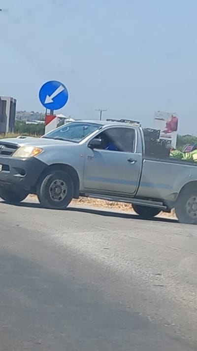A silver Toyota Hilux pickup truck is parked on the side of a road in Pyli, Greece. The truck has a single cab and a short bed. The bed of the truck is loaded with various items, including what appears to be green bags and possibly some fabric. A person is visible in the driver's seat, wearing a blue shirt. In the background, a blue circular traffic sign with a white arrow pointing left is mounted on a pole. The sky is clear and the sun is shining. The road appears to be paved.