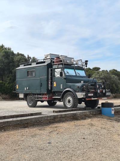 An olive-green, boxy camper truck is parked on a low concrete platform, with a gravel ground surrounding it. The truck is heavily modified, with a large front bumper, additional lights, and roof-mounted equipment. The camper section is a windowed, enclosed box structure. The license plate of the truck reads "HH-H5665H". Trees and other greenery are visible in the background. The location is El Perelló, Spain, as suggested by the user. A blue and black cylindrical object is present near the front of the truck.